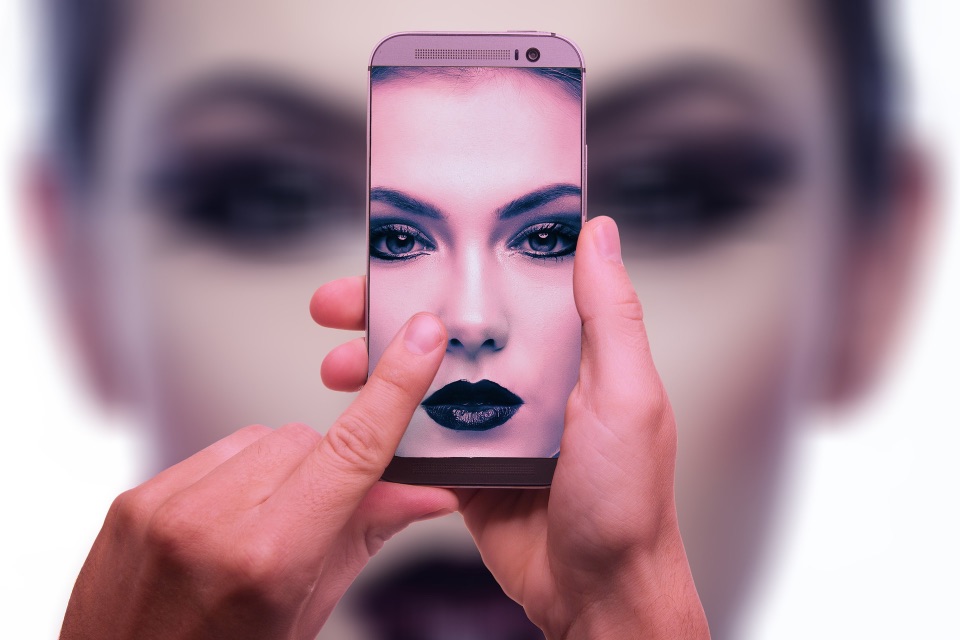 Bild einer in dunklen Tönen geschminkten Frau, die ein Handy im Selfiemodus hält. Darauf ist ihr düster dreinschauendes Gesicht zu sehen.
