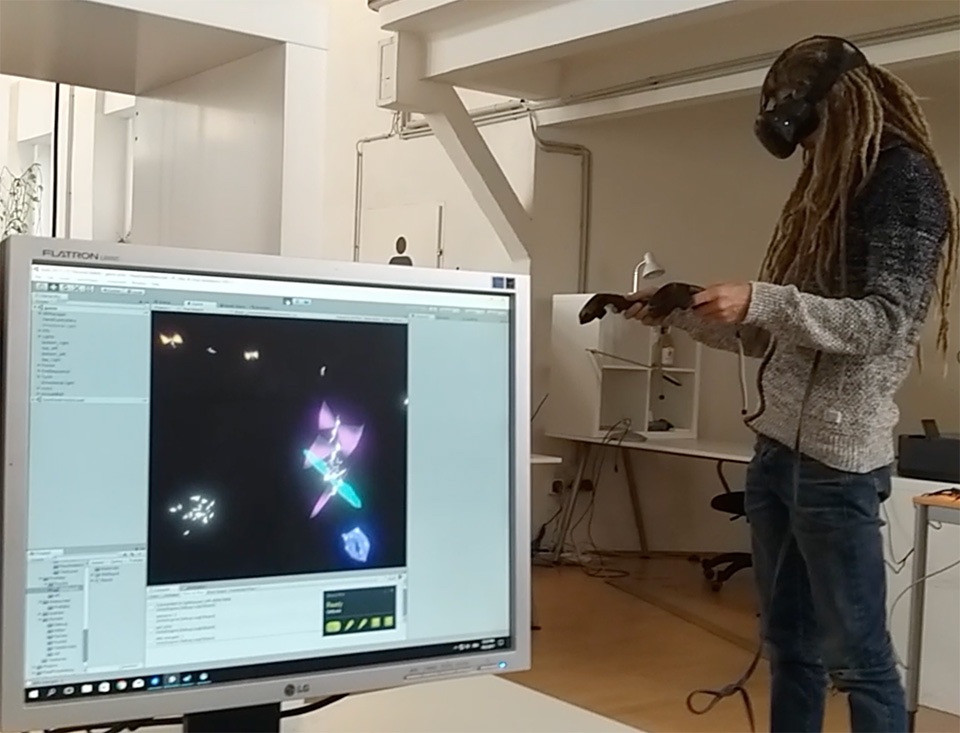 Der 3D-Animator Tom Stockinger testet mit VR-Brille und Controllern in einem Büro seine 3D-Modelle der Feen, die man im Vordergrund auf einem Computerbildschirm sehen kann. 