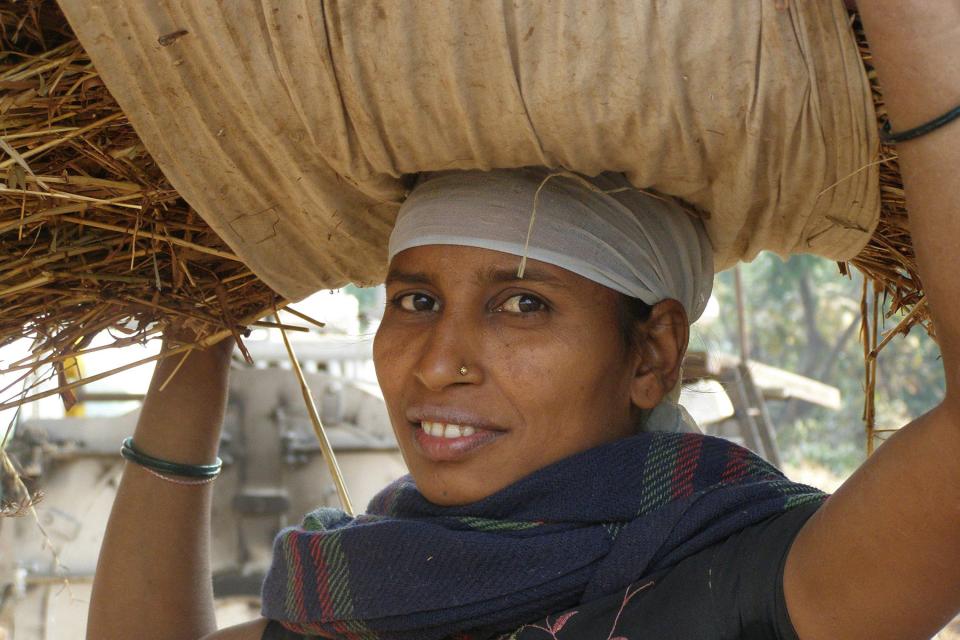 Eine Frau mit weißen Kopftuch trägt einen großen Heuballen am Kopf und sieht dabei lächelnd in die Kamera, Trägerin aus Afrika