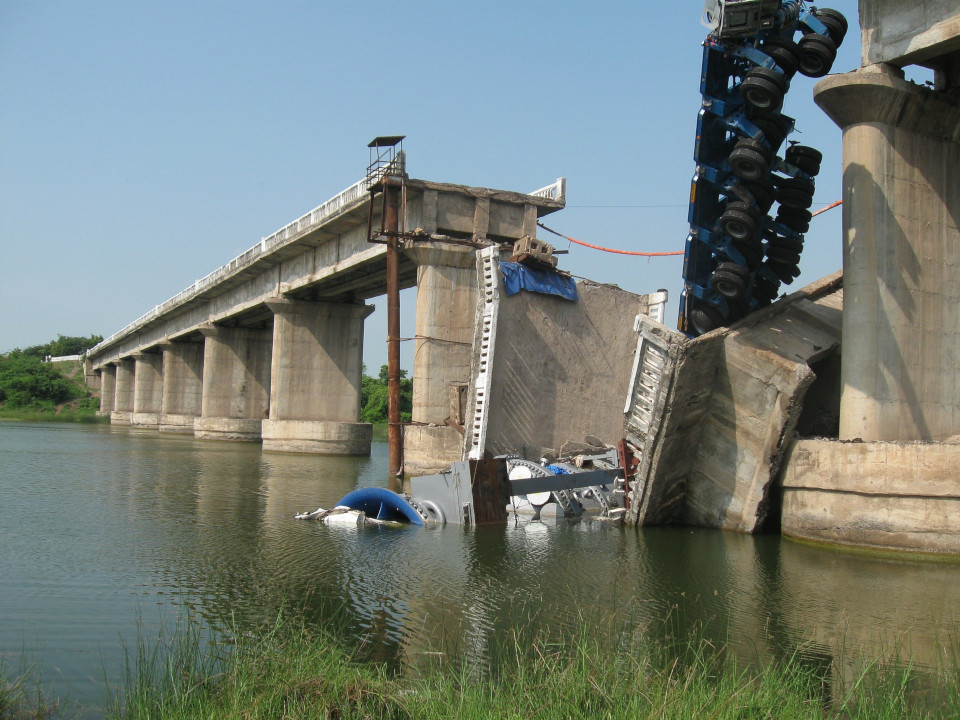 Bild einer  Betonbrücke über einem Fluss, wo ein Abschnitt zwischen zwei Pfahlen eingestürzt ist und ein nicht zuordenbares Fahrzeug mit in den Trümmern liegt.