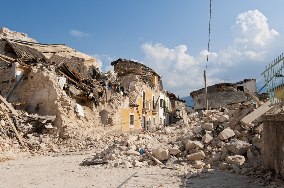 Foto einer Häuserruine nach einem Erdbeben, nur noch eine gelbe Hausfassade steht, alles andere liegt in Form von großen Steinbrocken auf dem Weg verteilt, im Hintergrund ein blauer Himmel.