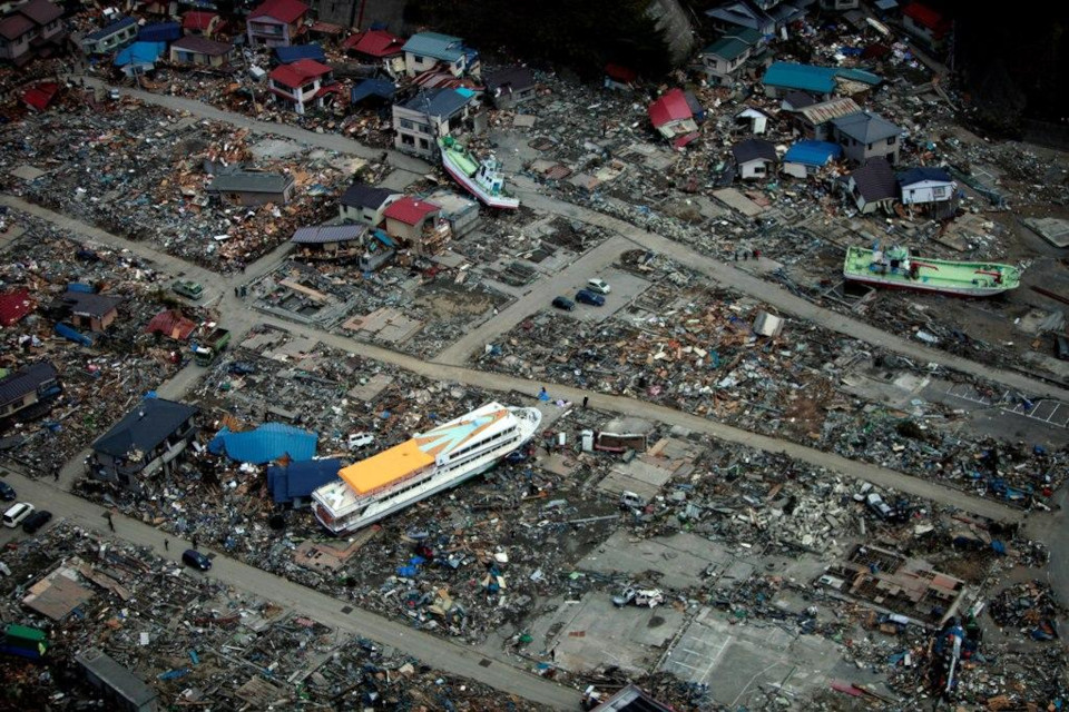 Dronenbild von ins Inland angespühlten Booten und einer gelben Fähre, welche zwischen zerstörten Häusertrümmern liegen, nach einem Tsunami in Japan.