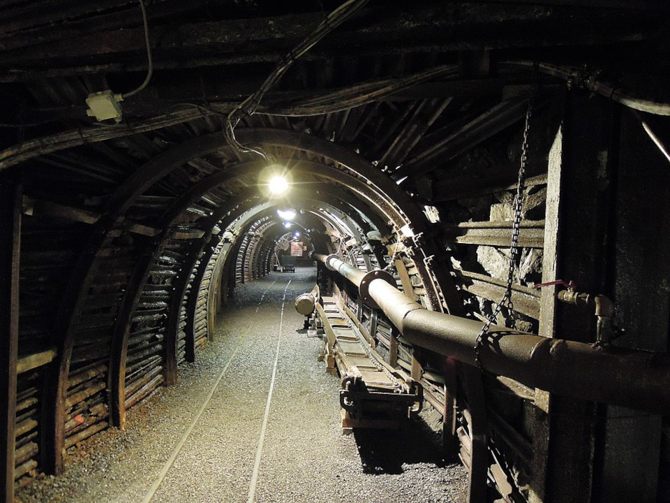Foto von einem menschenleeren düsteren Bergerwerkstollen. Ein dunkler niedriger Tunnel, mit grellen Glühbirnen bzw. Lichtern an der Decke und einem großen Rohr rechts entlang der Wand. 