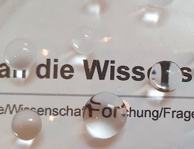 Wassertropfen auf einer Glasscheibe. Dahinter sieht man ein Zeitung mit dem Wort „Wissenschaft“ durchscheinen.